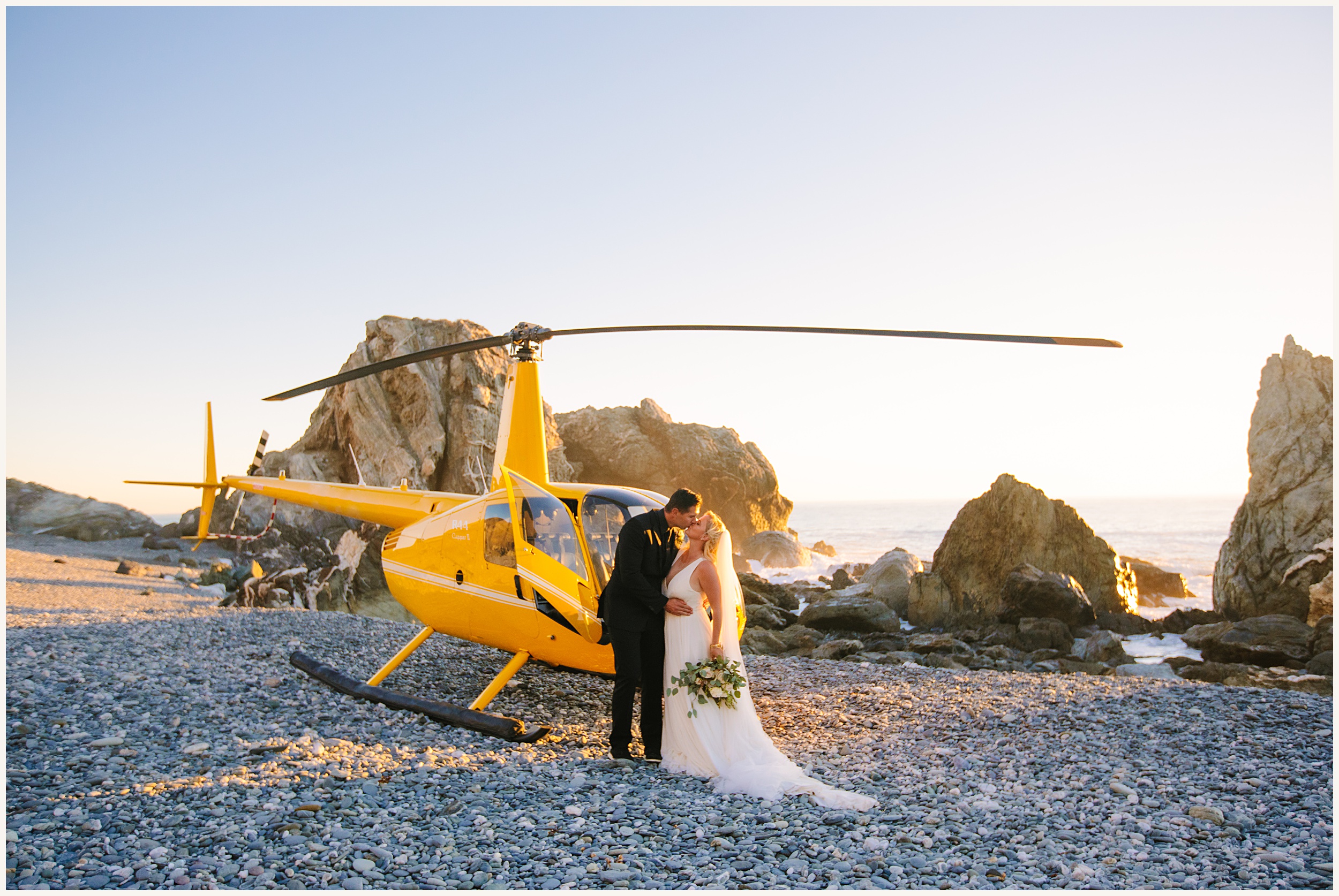 Photo of Bride and groom on rock beach in front of yellow helicopter