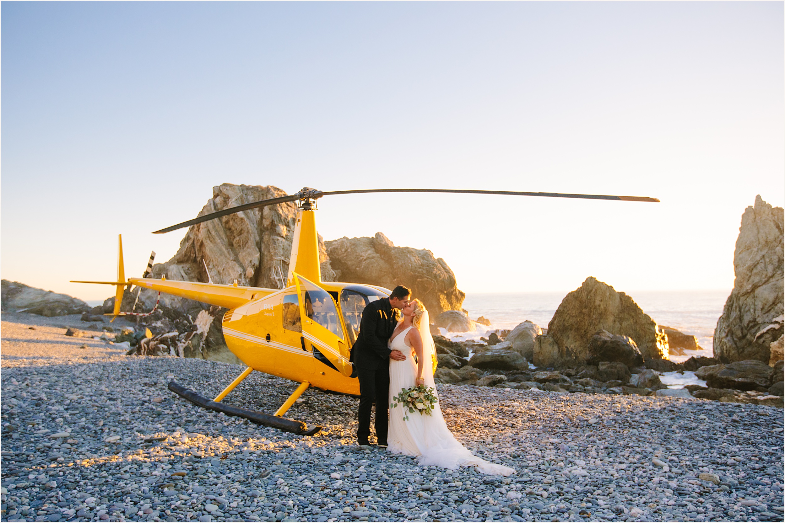 bride and groom standing on a rock beach kissing and standing in front of a yellow helicopter