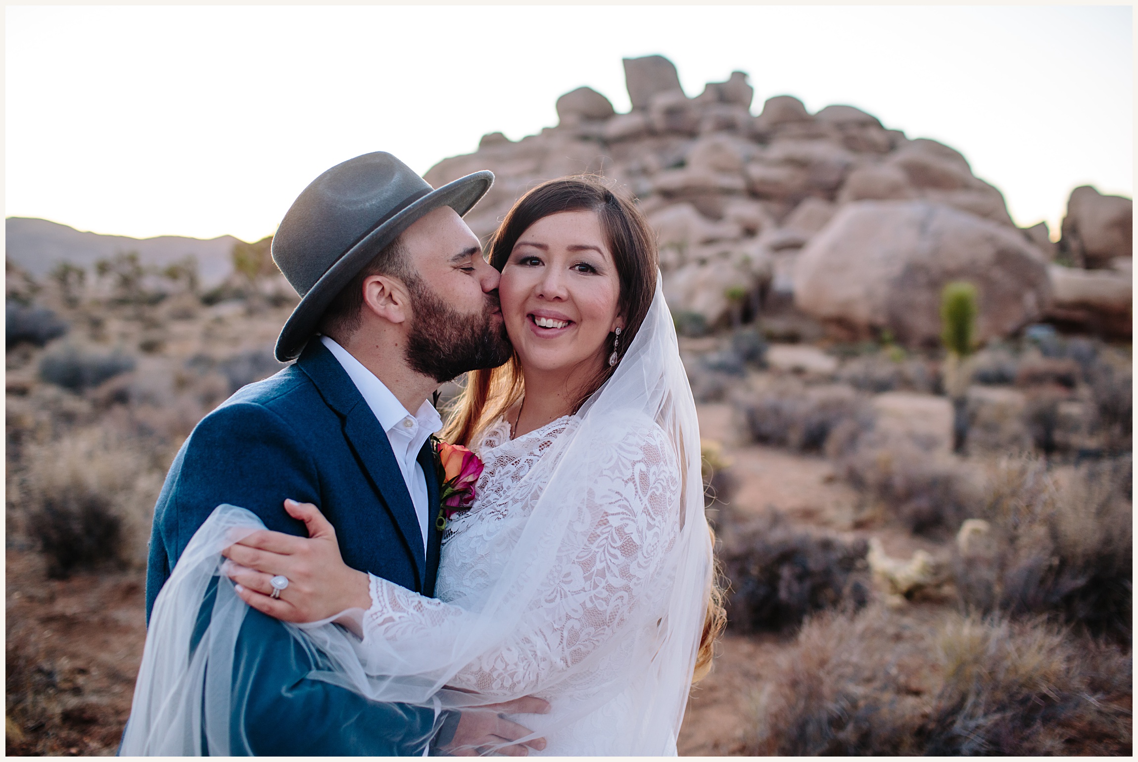 Photo of the groom in a blue suit and grey fedora kissing brides cheek who is in a elegant white long sleeve elopement dress