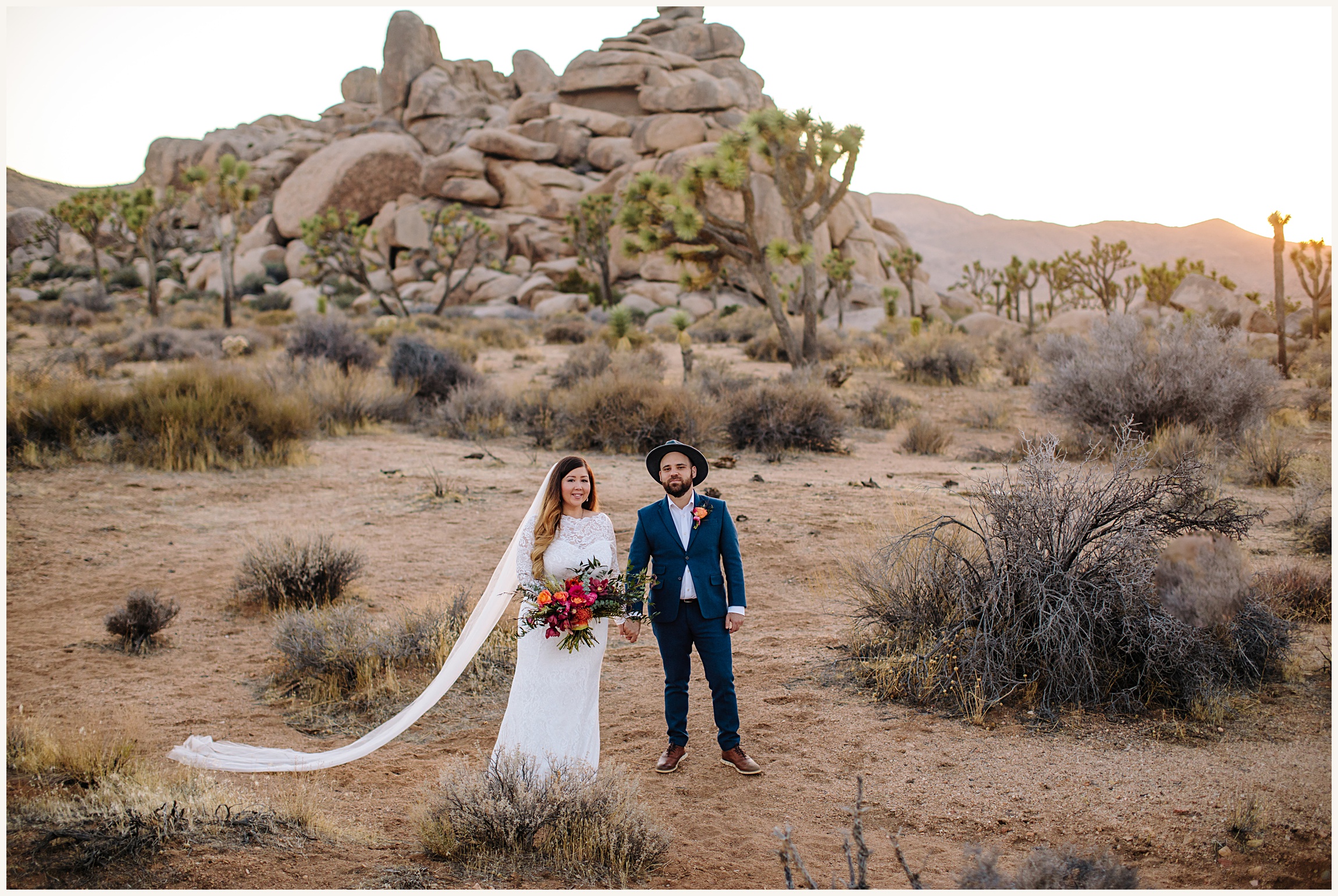 Photo of bride in and elegant white lace long sleeve elopement dress holding a multicolored pink bouquet and groom in a blue wedding suit with a fedora at Joshau Tree