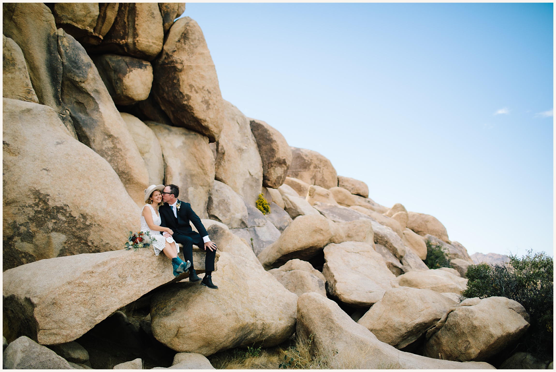 Photo of bride wearing an elegant white lace A-line elopement dress and groom wearing a black wedding suit and white button up on Joshua tree rocks