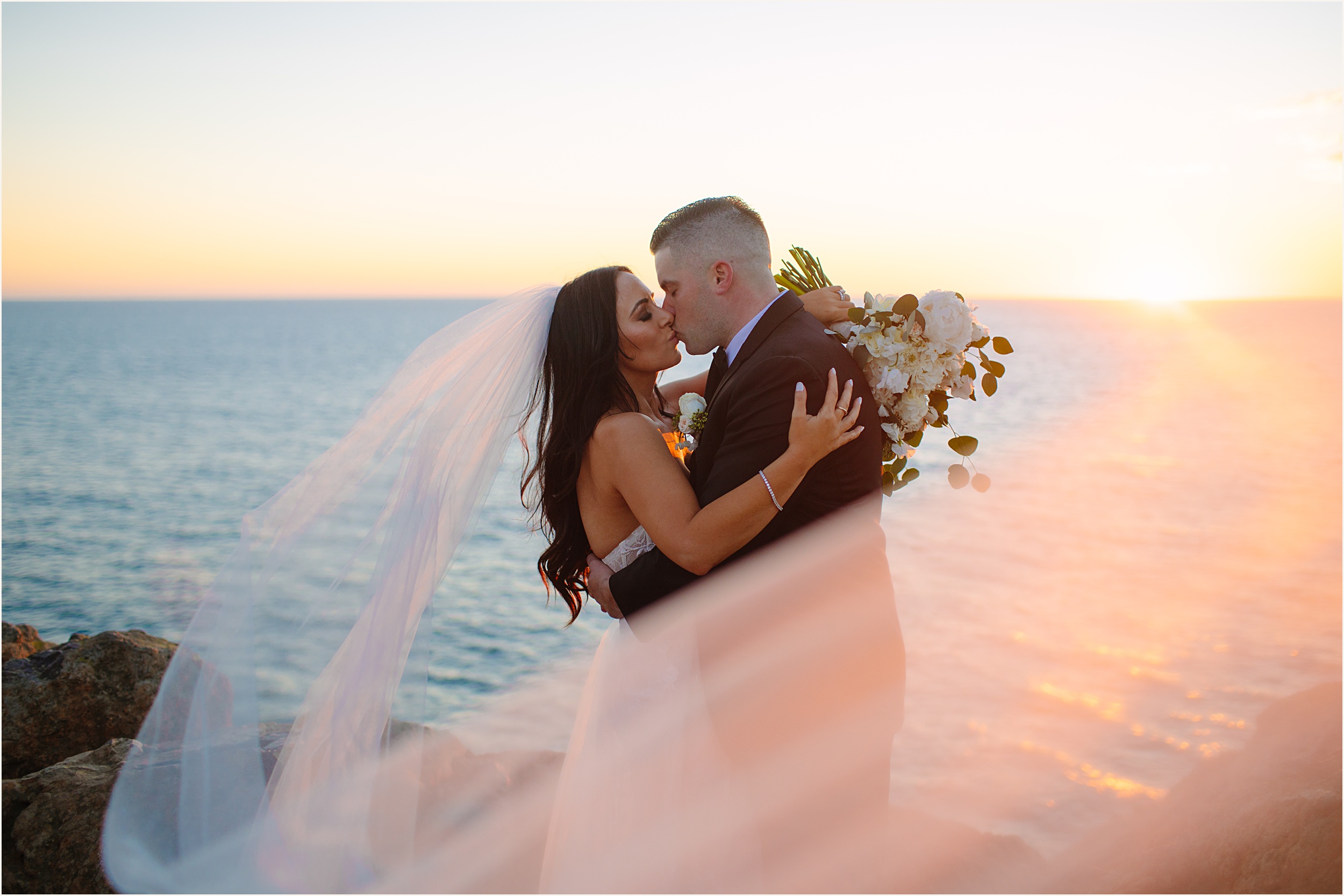 Photo of bride and groom kissing on cliffside view with sun setting