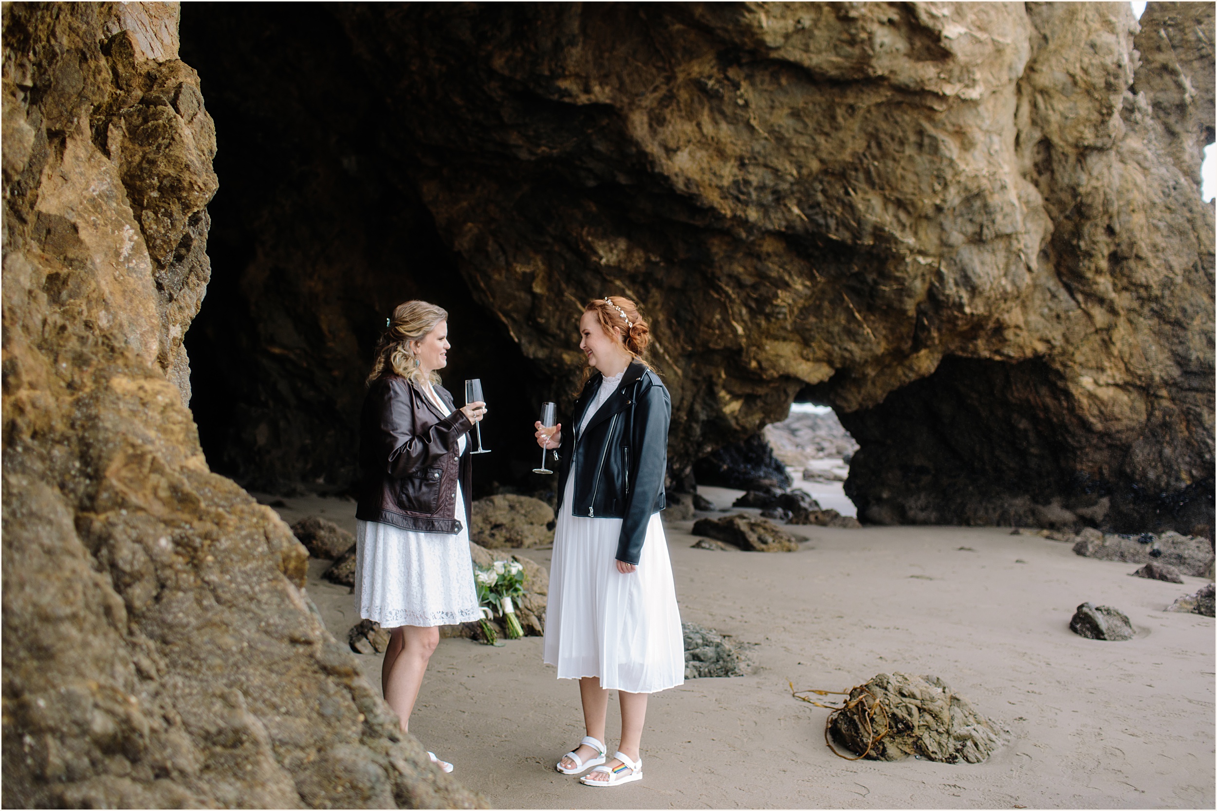 Photo of brides champagne toasting in stunning white lace elopement dresses and leather jackets at El Matador beach