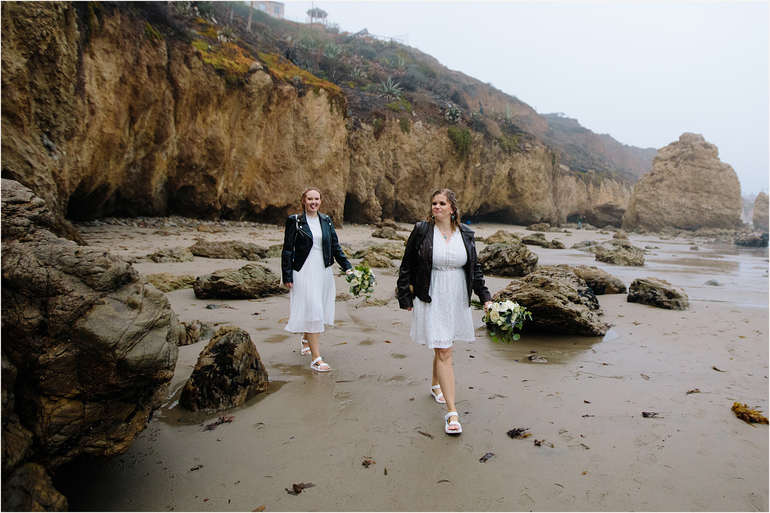 Photo of brides walking down El Matador beach in stunning white lace elopement dresses and leather jackets