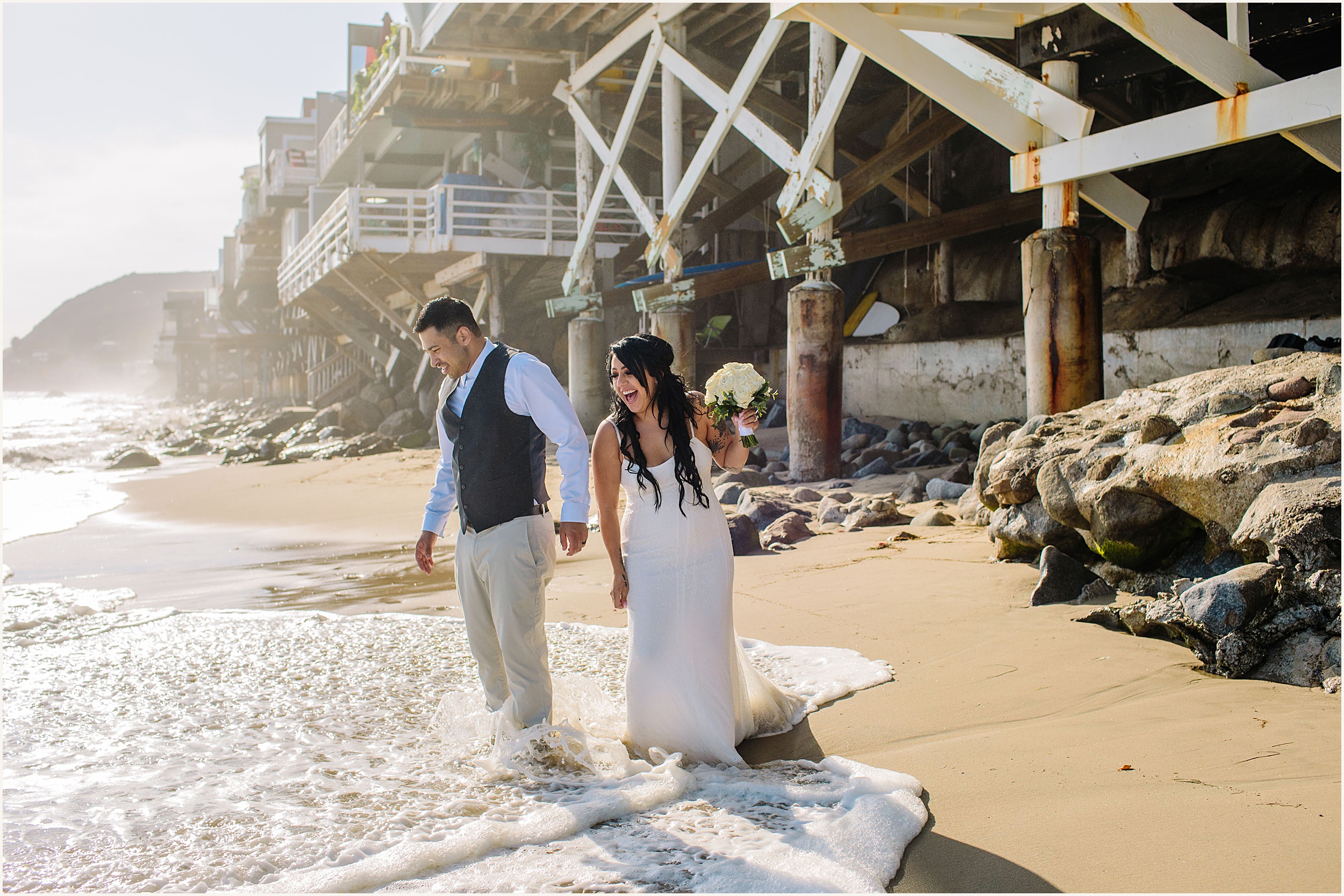 bride and groom stand on beach as the wave splashes their feet